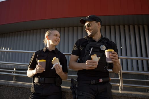 Male and female patrol police officers enjoying lunch break. Policeman and policewoman resting outdoor, drinking coffee, eating sandwich and having friendly conversation