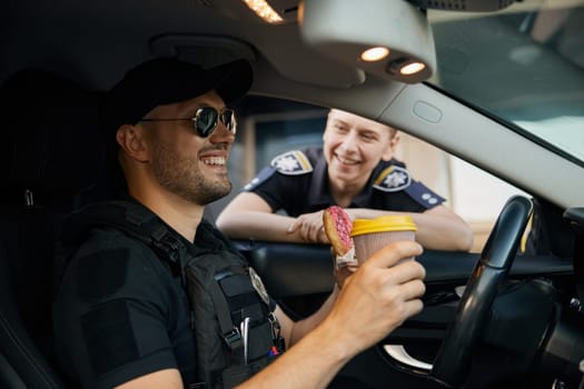Smiling patrol police officers diverse team sitting in car having coffee break time at work. Policeman with donut and hot aroma drink