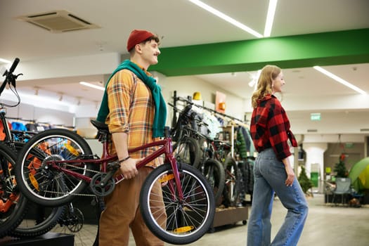 Happy man and woman carrying new kids bike walking through sports shop showcase. Married couple buying new bicycle for their child as birthday gift