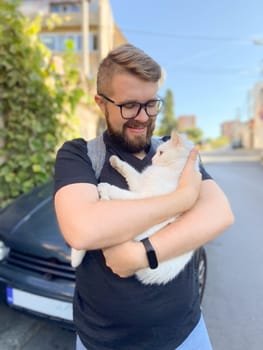 Bearded man gently embraces a white cat, conveying a moment of warmth and connection with pet. Outdoor setting adds an element of everyday life and companionship.