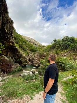 Millennial man stands thoughtfully by a stream in a rocky, verdant canyon, taking in the tranquility of the nature surrounding him. Solitude and the meditative beauty of an untouched landscape.