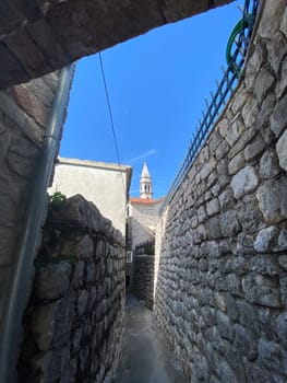 Narrow stone alleyway leading to a historic tower against a clear blue sky, capturing charm and mystery of ancient architecture. Confined passage and towering structures evoke a sense of exploration and timeless beauty.