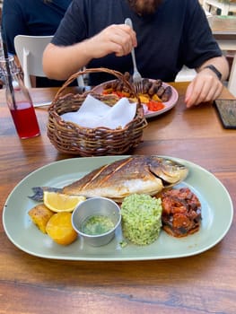 Delicious plate featuring a grilled fish with sides of rice and vegetables sits on a wooden table, ready for a delightful meal. Person in background enjoys their food, creating a warm and inviting dining scene.