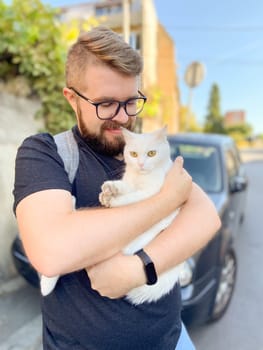 Bearded man gently embraces a white cat, conveying a moment of warmth and connection with pet. Outdoor setting adds an element of everyday life and companionship.