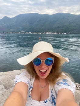Funny woman in a sun hat and sunglasses smiles joyfully by a serene lake with majestic mountains in background. Taking selfie with moment of happiness and natural beauty during her vacation.