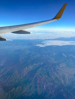 View of airplane wing through window at high altitude, showcasing clear blue sky and clouds far below. Serene beauty of flight and the expansive view of the horizon from above.