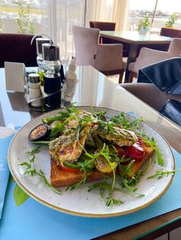 Close-up plate of grilled vegetables and chicken on toast is served in a stylish, bright seaside restaurant. Dish is garnished with fresh arugula and accompanied by a variety of seasonings and condiments on table.