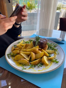 Close-up man takes a photo of a dish consisting of grilled meat and potatoes. Social networks, blog and food blogger.
