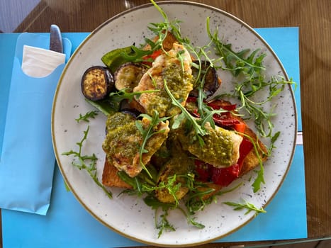 Close-up plate of grilled vegetables and chicken on toast is served in a stylish, bright seaside restaurant. Dish is garnished with fresh arugula and accompanied by a variety of seasonings and condiments on table.
