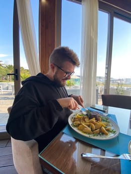 Bearded man enjoys a meal at a seaside restaurant, with a plate of steak and potatoes. Bright and open atmosphere, surrounded by palm trees and a scenic view of the beach outside.
