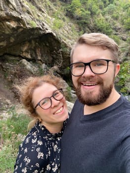 Millennial joyful couple takes a selfie in a lush, mountainous landscape, celebrating their outdoor adventure. Beauty of nature and the happiness of shared experiences.