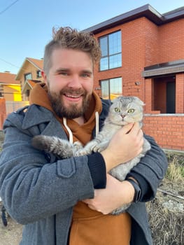 Millennial man in a coat holds a cream-colored Scottish Fold cat in his arms in autumn suburban street. Beautiful pet. Domestic cat