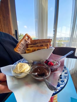 Close-up male holding a plate of waffles with sides of ice cream, berries, and chocolate at a seaside restaurant. Bright and sunny setting, with palm trees visible outside, creates a relaxed vacation mood.