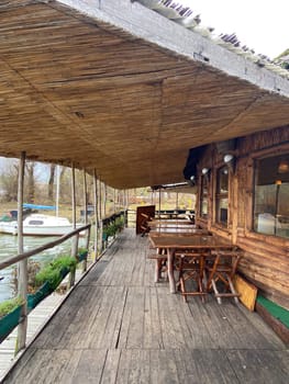 Rustic waterfront deck with wooden tables and a thatched roof overlooks serene river. Outdoor seating area offers a tranquil place to relax and enjoy the natural surroundings.