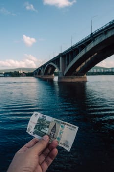 Hand with 10 rubles bill on background of Communal bridge in Krasnoyarsk through the Yenisei River in Russia, Krasnoyarsk. This bridge is depicted on the 10 ruble bill