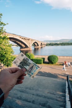 Hand with 10 rubles bill on background of Communal bridge in Krasnoyarsk through the Yenisei River in Russia, Krasnoyarsk. This bridge is depicted on the 10 ruble bill