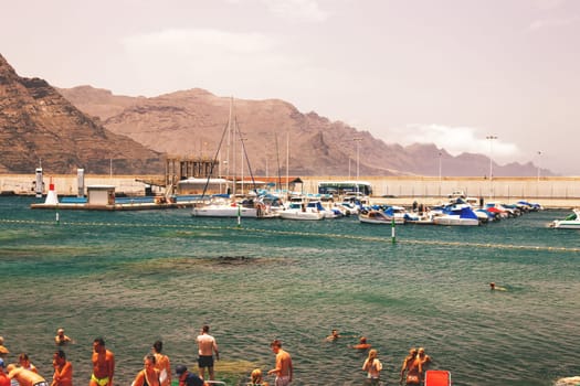 Puerto de las Nieves, Gran Canaria Island, Spain - 20 July 2024. Tourists on the stony beach Playa de las Nieves with a view of the harbour.