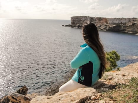 Woman sitting on a cliff gazing at the ocean under a sunny sky, latin woman looking out over the mediterranean sea cliffs, in mallorca balearic islands spain