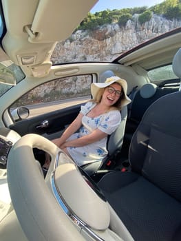 Cheerful woman is sitting in a car with the roof open, enjoying a sunny day. Wearing a summer hat and laughing with joy.