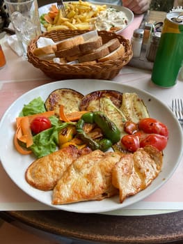 Delicious meal featuring grilled vegetables and chicken breast is elegantly presented on a white plate. Basket of sliced bread and a soft drink can, creating a cozy dining atmosphere.