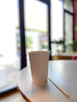 Simple white paper cup sits on a round table in a cozy cafe, inviting a moment of relaxation and warmth. Blurred background suggests a peaceful indoor setting, perfect for enjoying a quiet coffee break