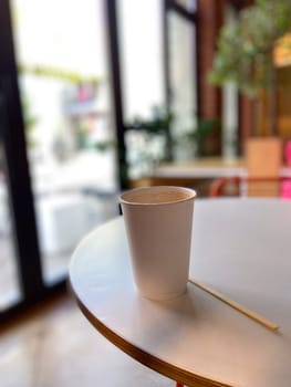 Simple white paper cup sits on a round table in a cozy cafe, inviting a moment of relaxation and warmth. Blurred background suggests a peaceful indoor setting, perfect for enjoying a quiet coffee break