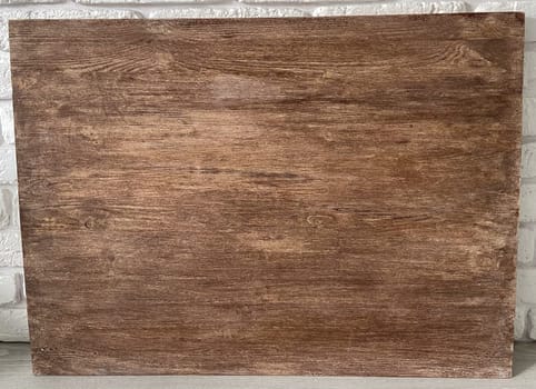 Rustic wooden panel leans gracefully against a textured white brick wall, showcasing its rich grain patterns and natural warmth. Contrast between the organic wood and the structured brick provides a harmonious blend of nature and architecture.