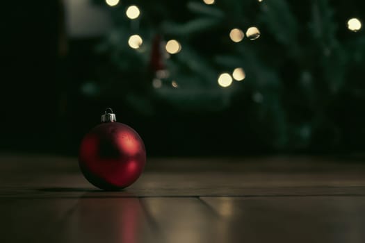 A solitary red ornament rests on the wooden floor illuminated by soft lights from a nearby Christmas tree.