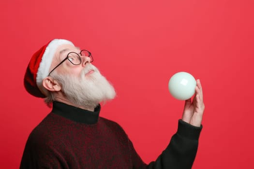 A joyful man dressed as Santa Claus gazes upward while balancing a Christmas ornament in his hand set against a vibrant red background.