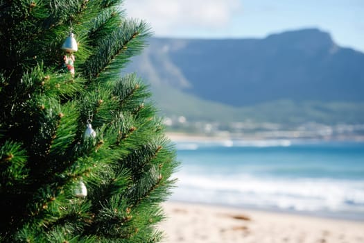 A decorated Christmas tree stands on the sandy beach with waves crashing and a mountain backdrop in Cape Town under clear skies.