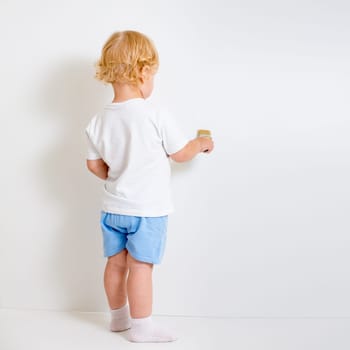 Baby boy with paint brush rear view standing near blank white wall