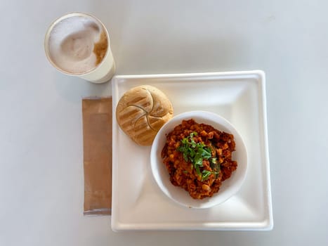 Delicious meal featuring a hearty bowl of stew garnished with herbs, served alongside fresh bread and a frothy coffee, is presented on a clean white tray. Inviting combination of flavors and textures perfect for a satisfying breakfast or lunch.