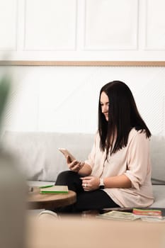 A brunette girl uses a mobile phone app, plays games, makes purchases online, orders delivery, relaxes on the couch.