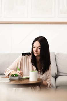 A brunette girl sitting at a desk in the office, making notes in a notebook on work with a mug of tea