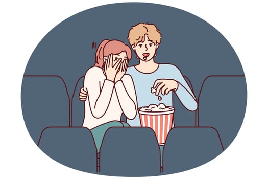 Guy and girl are watching horror movie in cinema and eating popcorn enjoying films about vampires. Fearful woman closes eyes with hands sitting in cinema near man watching horror movie with interest