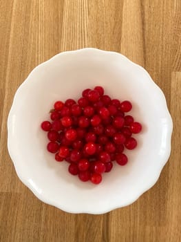 Collection of vibrant red berries sits elegantly in a white bowl, creating a striking contrast against a wooden background. Simple composition showcases the freshness and natural appeal of the fruit.