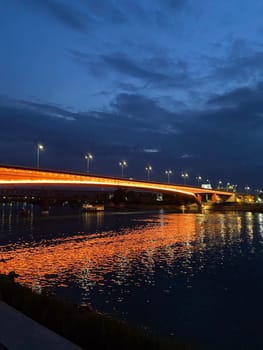 Vibrant bridge illuminated with orange lights stretches across a tranquil river at dusk, reflecting beautifully on the water surface. Deep blue sky provides a stunning contrast, creating a captivating and serene evening spectacle.
