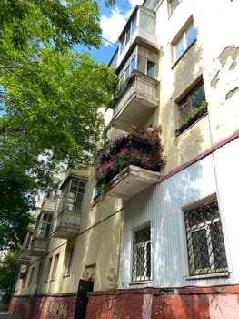Vibrant urban apartment building stands tall, with colorful flowers adorning its balconies against a backdrop of lush green trees and a bright blue sky. Harmonious blend of nature and city life, highlighting a touch of beauty in everyday surroundings.