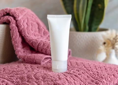 White cream squeeze tube on pink folded towels near green plant in bathroom, close up, cosmetic packaging mockup. Natural mens face cream for personal care
