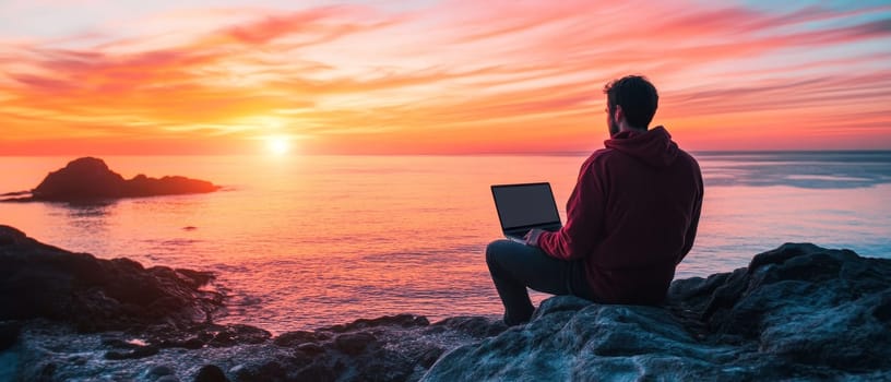 Silhouette of person working on laptop while sitting on rocky shore, against a breathtaking orange and pink sunset over calm sea.