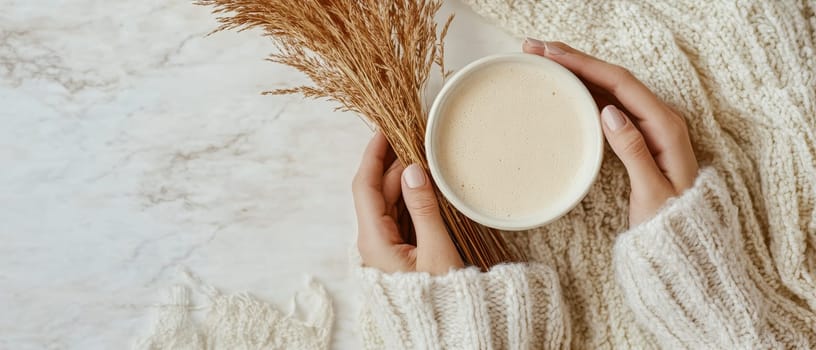 Hands holding warm coffee mug with dried wheat stalks, against soft knit sweater background, evoking comfort and autumn coziness.
