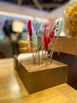 Vibrant arrangement of dried flowers in a wooden holder adds a pop of color to a cozy restaurant setting, with twinkling lights in the background. The mix of red and blue blooms against a warm, soft focus creates a festive and inviting atmosphere.