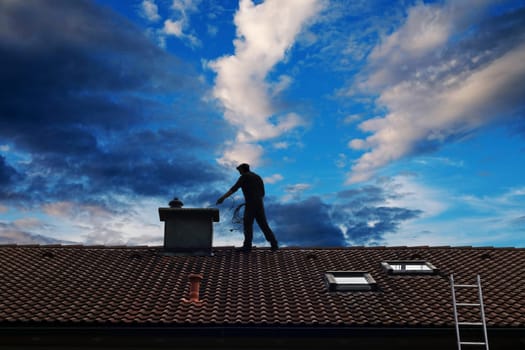 A chimney sweep cleans the chimney on the roof of a house in front of a beautiful sky