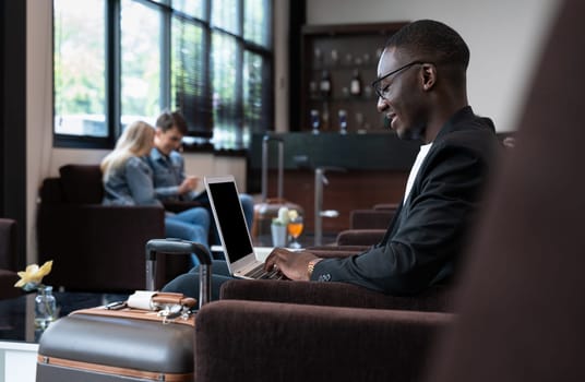 African businessman using laptop while sitting in airport lounge with blurred couple in background. Business travelers waiting for their flight at airport terminal.