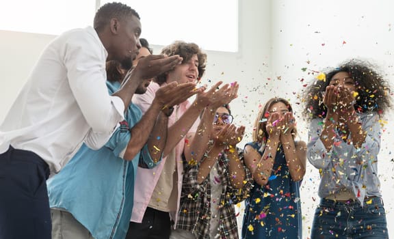 Group of six people friends in casual dress standing close together, celebrating party and blowing colorful confetti into air. Multicultural friends having fun together on weekend at home.