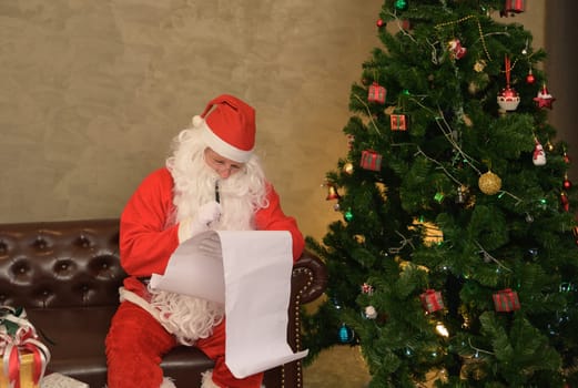Santa Claus sitting on leather couch and reading long list for giving Christmas presents to child. Man dressed in red suit and hat at home by decorated Christmas tree with colorful ornaments and twinkling lights. Christmas and New Year concept.
