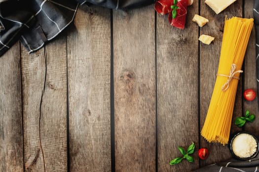 Italian cuisine concept flat lay. Parmesan Cheese chunks, grated cheese in black bowl, raw pasta, thinly sliced ham, cuts of cherry tomatos, basil leaves branches, kitchen towel on wooden background