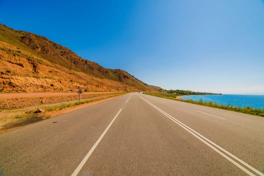 asphalt interstate road between yellow sedimentary rock mountain and large lake at sunny summer day, wide angle view
