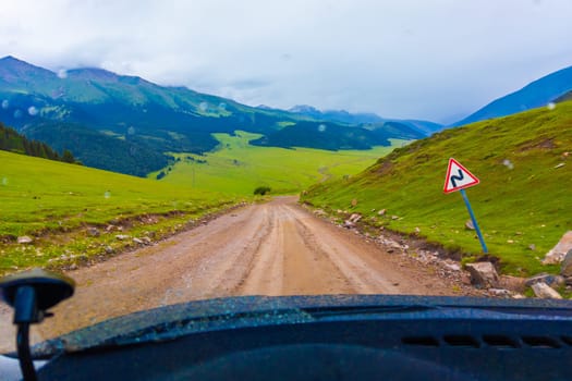driving along the dirt road at rainy summer day in mountains with spruce forest on mountain slope, view from driver perspective