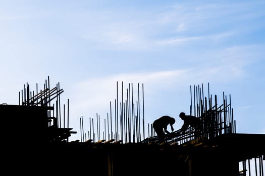 unrecognizable workers are working at construction site with many vertical rebars building a structure in front of blue sky in silhouettes.